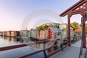 A wooden bridge spans the serene Nidelva River, flanked by iconic wooden houses bathed in the warm glow of a summer sunset in