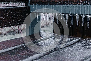 A wooden brown bench covers a layer of ice and icicles after freezing rain in winter