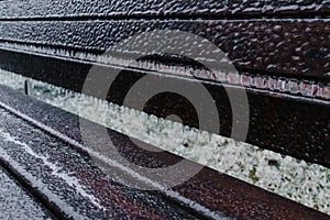 A wooden brown bench covers a layer of ice and icicles after freezing rain in winter