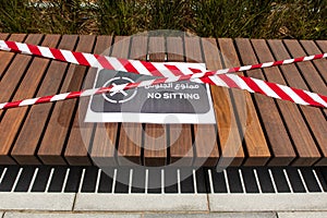 Wooden brown bench with `No Sitting` sign in english and arabic and red and white barricade tape, COVID-19 in Dubai