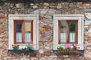 A wooden, brown windows and a fragment of a stone wall of the building. Curtains hanging in the window, colorful flowers