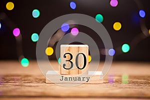 Wooden calendar with black 30 january word at center of dark background with garland bokeh. Winter time, year concept