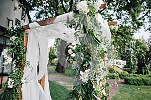 Wooden ceremony arch decoretade by white cloth, flowers and greenery standing in bright garden for wedding ceremony. Decor.