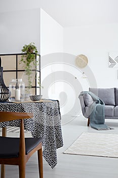 Wooden chair at dining table in white apartment interior with grey sofa with green blanket. Real photo