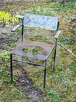 A wooden chair, a hole in the middle of the chair to fit a toilet, an old object