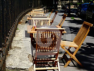 Wooden chairs, restaurant tables and terrace along old rusty wrought iron fence on street in bright light
