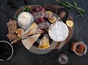 Wooden cheeseboard on slate surface with a variety of cheeses, crackers, fruit, honey, rosemary sprigs and chutney