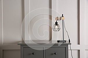 Wooden chest of drawers with modern lamp near white wall, space for text. Interior element