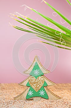 Wooden Christmas tree with sparkles in the sand with a palm tree on a pink background. Holidays in hot countries. Vertical