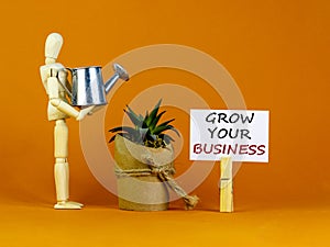 Wooden clothespin with white sheet of paper. Text `grow your business`. Wooden model of a human watering a house plant from a