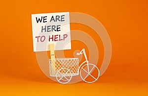 Wooden clothespin with white sheet of paper. Text `we are here to help`. Miniature bicycle model. Beautiful orange background.