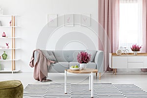 Wooden coffee table with fresh heather and fruits on plate in real photo of bright sitting room interior with posters on wall