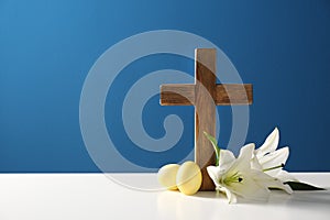 Wooden cross, Easter eggs and blossom lilies on table against color background