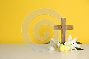 Wooden cross, Easter eggs and blossom lilies on table against color background