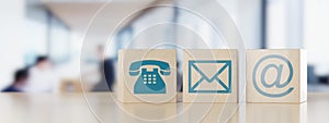 Wooden cube or box telephone, envelope letter and e-mail symbols in a row on wooden table with a blurred office in the background