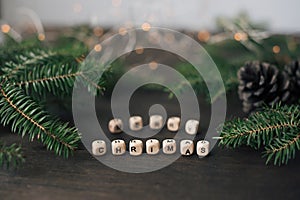Wooden cubes with letters make up the phrase Merry Christmas on a wooden table. Fir branches, cones and a garland in the
