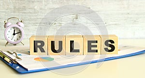 Wooden cubes lie on a folder with financial charts on a gray background. The cubes make up the word RULES.