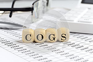 Wooden cubes with the word COGS on a financial background with chart, calculator, pen and glasses, business concept