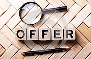 Wooden cubes with the word OFFER stand on a wooden background between a magnifying glass and a pen