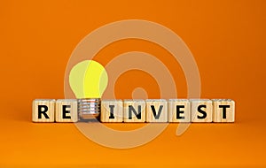 Wooden cubes with word `reinvest`. Yellow light bulb. Beautiful orange background. Business concept. Copy space