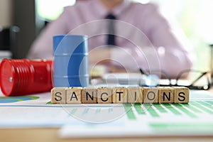 Wooden cubes with word sanction, oil drums and documents on table and msnager in background.