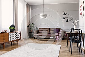 Wooden cupboard in bright living room interior with leather sofa and chairs at table. Real photo