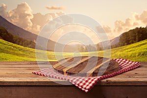 Wooden cutting board with checked tablecloth on table over landscape background