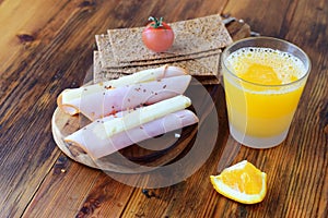 Wooden cutting board with crispbread, cheese, ham, cherry tomato, glass of fresh orange juice on a wooden background