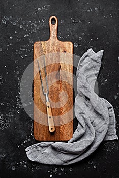 Wooden cutting board  meat fork and napkin on black stone background. Kitchenware. Top view  flat lay