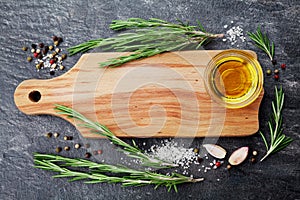 Wooden cutting board, olive oil, rosemary plant, salt, garlic and pepper on black table from above for food cooking background or