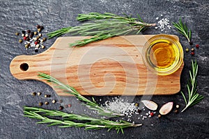 Wooden cutting board, olive oil, rosemary plant, salt, garlic and pepper on black table from above for food cooking background or photo