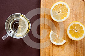 On a wooden cutting Board, slices of lemon and a transparent Cup with lemon on a brown background, top view