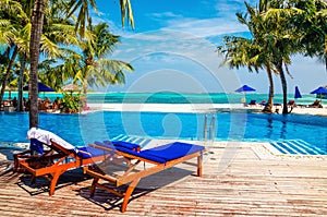 Wooden deck chairs with blue pillows above the pool. In the background, the azure water of the ocean and a beach with