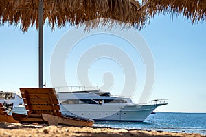 Wooden deck chairs under rough straw sun umbrella on sea beach and big white yacht ship in water near shore on sunny summer day