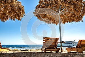 Wooden deck chairs under rough straw sun umbrella on sea beach and big white yacht ship in water near shore on sunny summer day