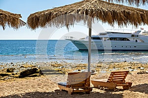 Wooden deck chairs under rough straw sun umbrella on sea beach and big white yacht ship in water near shore on sunny summer day