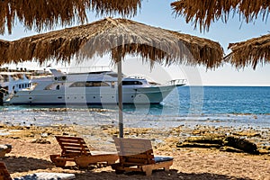 Wooden deck chairs under rough straw sun umbrella on sea beach and big white yacht ship in water near shore on sunny summer day