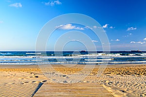 Wooden deck over sandy beach with blue sky and ocean on background. White foam on top of the ocean waves in Tarragona Spain