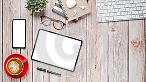 Wooden desk workplace with blank screen tablet and smartphone, keyboard, pen, eyeglass, notebook and cup of coffee,