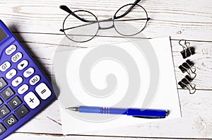 On the wooden desktop is a blue calculator, paper clips, glasses, a blue pen and an empty notepad with space to insert text. Copy