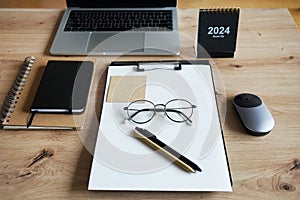 Wooden desktop with notepads, folder with papers, pens, stickers, glasses, laptop with mouse and calendar