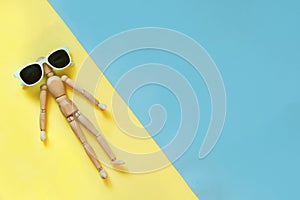 Wooden doll with sunglasses on yellow as sand beach and blue as water. Concept tan and beach. Danger of sunburn. Uv protec