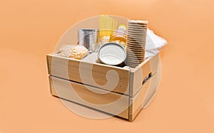 Wooden donation box with food: canned food, pasta, peas, millet, sugar, vegetable oil, cookies on an orange background.