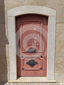 A wooden Door with faded and Peeling red paint, with highly decorated Metal Door Furniture set into a wall.