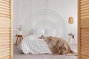 Wooden door and lamp in white bedroom interior with blanket on bed and flowers on table