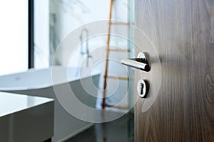 Wooden door open to nice modern bathroom, marble walls. selective focus to door handle, bathroom and bathtub blurred on background