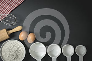 Wooden dough roller, a cup of flour, eggs and whisk on the black table