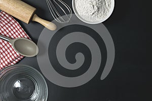 Wooden dough roller, a cup of flour and whisk on the black table