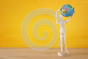 wooden dummy figure holding globe over yellow background. room for text.