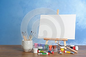 Wooden easel with blank canvas board and painting tools for children on table near color wall.